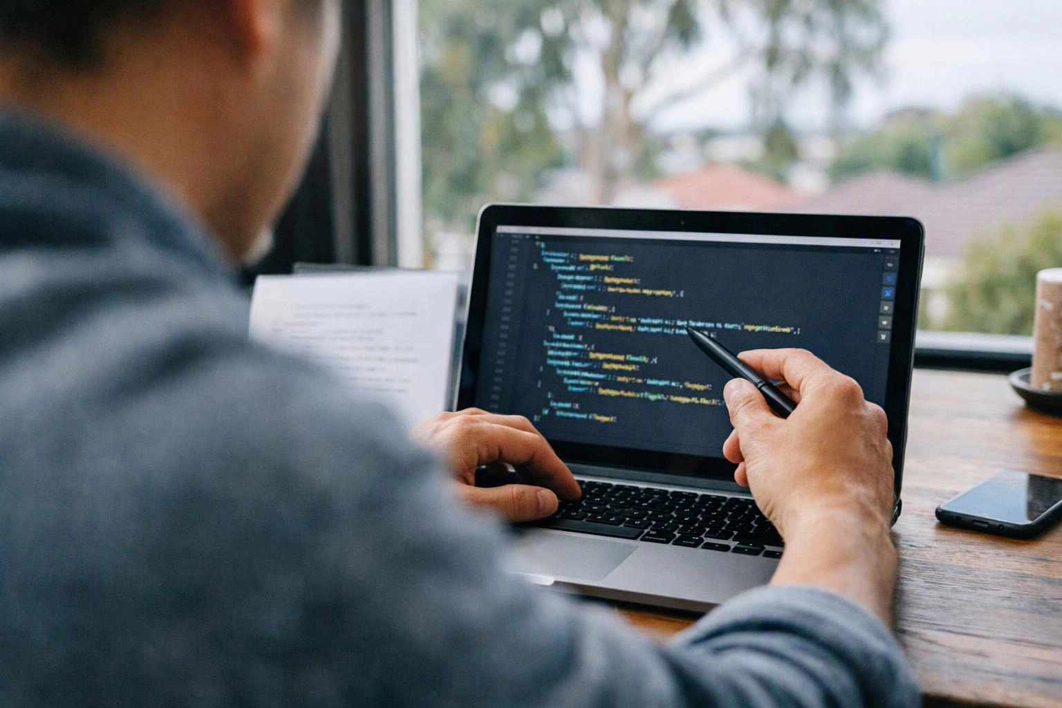 A person points at code on a laptop while sitting at a wooden table by a window