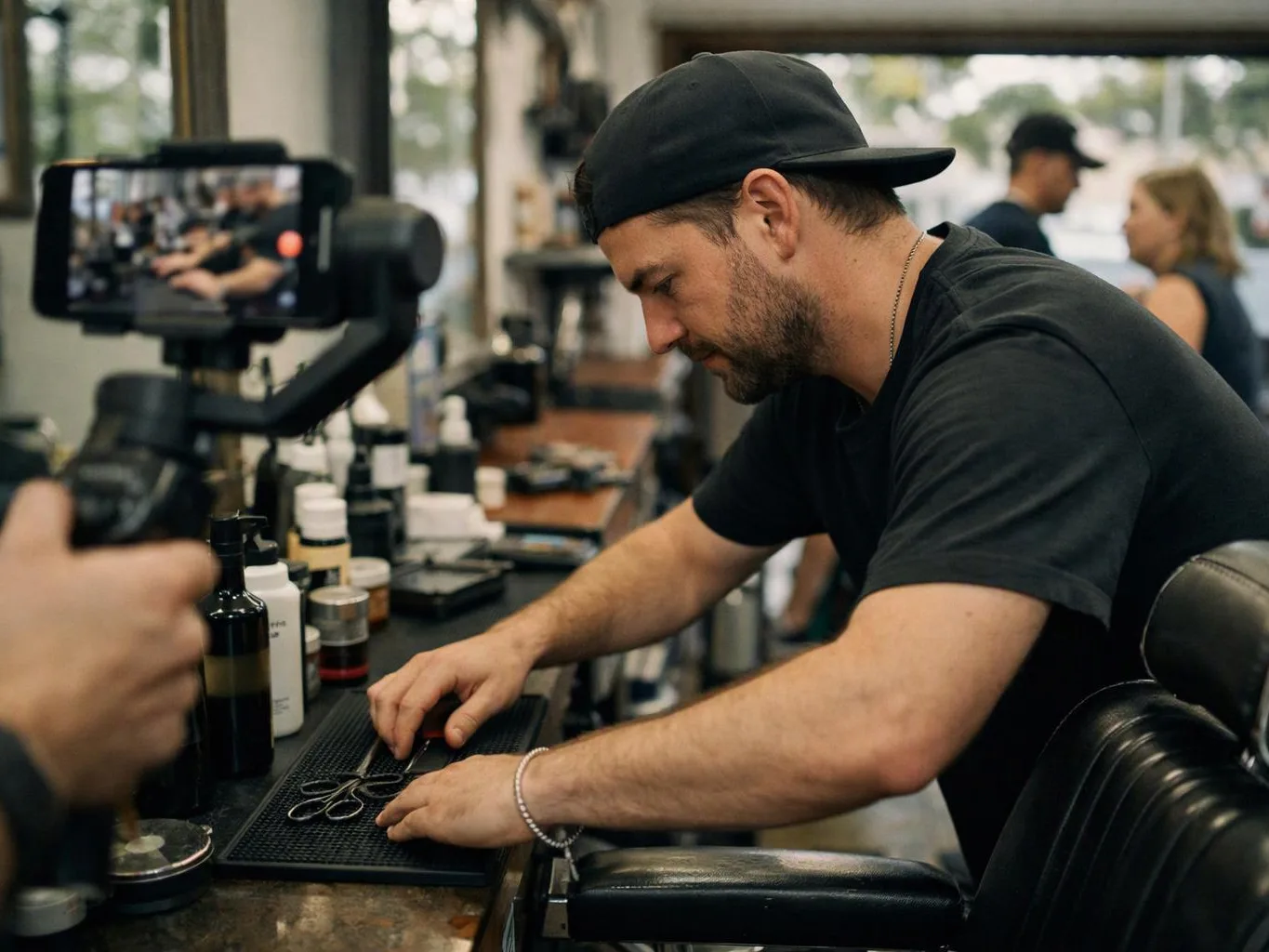 A man in a black cap trims tools at a barber station while a phone records nearby