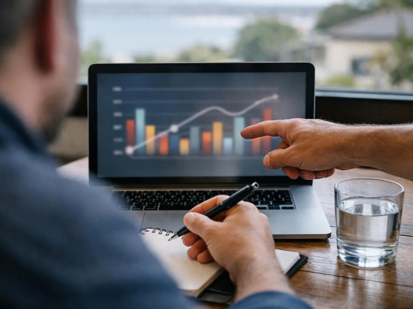 A person points at a laptop showing a colorful bar chart while another writes notes beside a glass of water